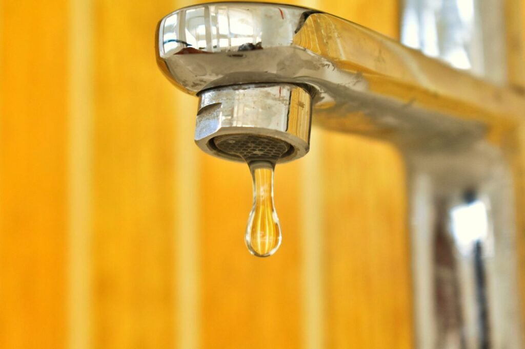 A close-up shot of a water droplet hanging from a stainless steel faucet, highlighting surface tension.