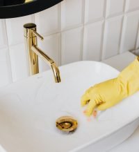 From above anonymous housekeeper in yellow rubber gloves wiping by sponge sink in bathroom